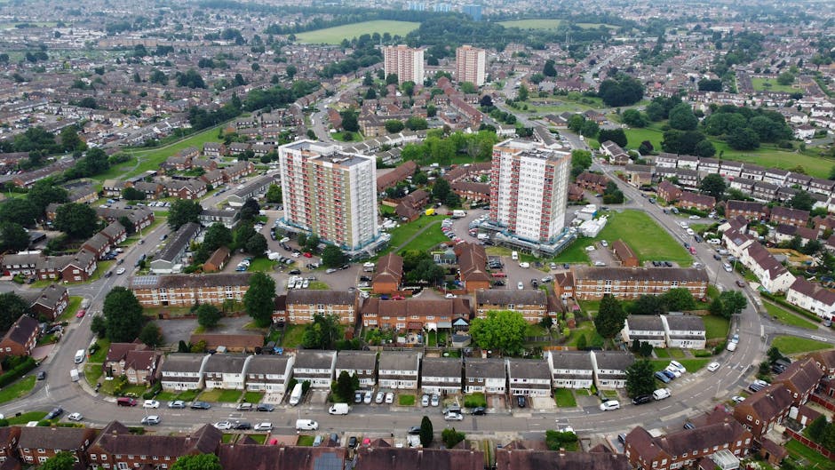 Aerial view of a residential neighbourhood in South Tottenham, showing a mix of low-rise terraced houses, smaller apartment buildings, and two taller high-rise apartment blocks with white exteriors and red accents. The area features numerous parked cars along curved streets, green spaces, and trees interspersed throughout the community. The image captures a typical urban scene relevant to house removals and furniture transport services, with the open spaces and road layouts indicating a well-organized environment suitable for home relocation and moving logistics. The lighting is natural with an overcast sky, highlighting the variety of building types and the overall layout of the neighbourhood as seen from above, underlining the context of local moving solutions provided by Man with Van South Tottenham.