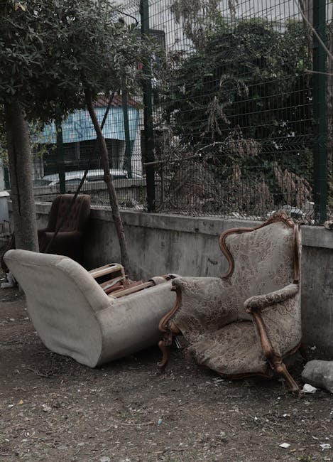 A photograph showing two antique upholstered armchairs placed outdoors on a dirt ground beside a concrete wall and metal fence. One armchair is covered in light-colored fabric with wooden arm supports and appears relatively intact, while the other has faded, worn upholstery with visible tears and missing padding, exposing the frame. The chairs are positioned close together, with one slightly tilted. In the background, there are trees, a metal fence, and a glimpse of buildings or structures behind the fence. The scene suggests furniture abandonment, possibly associated with home relocation or bulky waste disposal, and is related to services like furniture transport or removals provided by Man with Van South Tottenham.