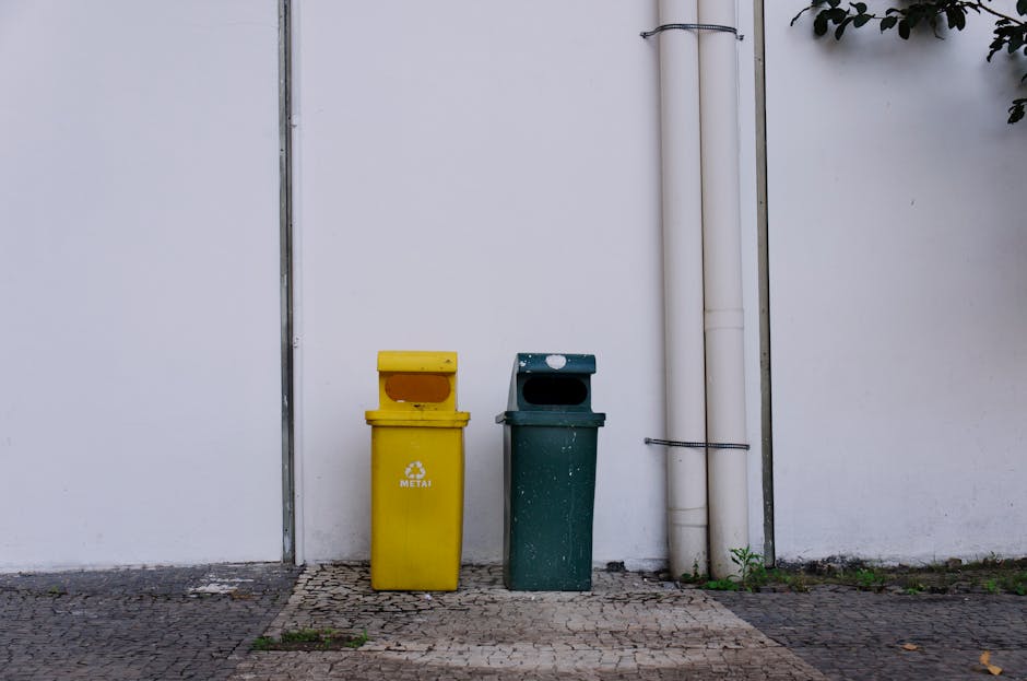 Two metal recycling bins, one yellow and one dark green, are positioned against a white exterior wall near a doorway in this outdoor scene. The yellow bin, which is marked with a recycling symbol and the word 'METAL', is placed to the left of the green bin, both standing on a paved surface made of small, textured bricks. To the right of the bins, two vertical white pipes run down the wall, secured with a black zip tie. The scene is illuminated by natural daylight, capturing a clean and organized setting typical of urban waste disposal or recycling collection areas, relevant to the logistics of house removals and moving services offered by Man with Van South Tottenham.