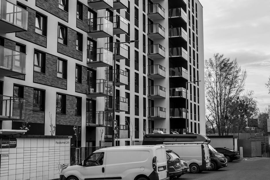 Black and white photograph of a modern multi-storey residential building with several balconies, some with glass railings, and large windows. In front of the building, a parking area contains several vehicles including a white van, a small black car, and a larger cargo van, all parked on a paved driveway. The vehicles are positioned near the building's entrance, which is partially visible. To the right of the vehicles, there are a few leafless trees and a small wall with a parking sign. The scene appears to depict the logistics of home relocation or furniture transport, with the vehicles ready for loading or unloading. The environment is illuminated by natural daylight, emphasizing the structure's contemporary architectural features, and subtly supports the context of moving and clearance processes associated with house removals in South Tottenham. Man with Van South Tottenham's service role in such relocation scenarios is implied through the setting.