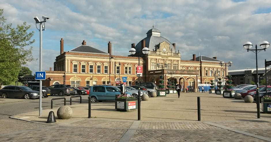 A spacious outdoor area in front of a historic brick building with ornate architectural details, including a central clock tower and decorative roofline, situated within a parking lot with multiple parked cars. The foreground features a paved area with large spherical stone bollards, metal bike racks, and a blue 'IN' sign indicating directions for vehicles. Several street lamps with spherical light fixtures are positioned along the perimeter, while flower planters and a few pedestrians are visible nearby. The scene is lit by daylight with a partly cloudy sky overhead. This setting illustrates the type of urban environment where house removals and furniture transport services, provided by Man with Van South Tottenham, are coordinated during home relocation or moving processes, ensuring safe loading of furniture and packing materials near residential and commercial premises.
