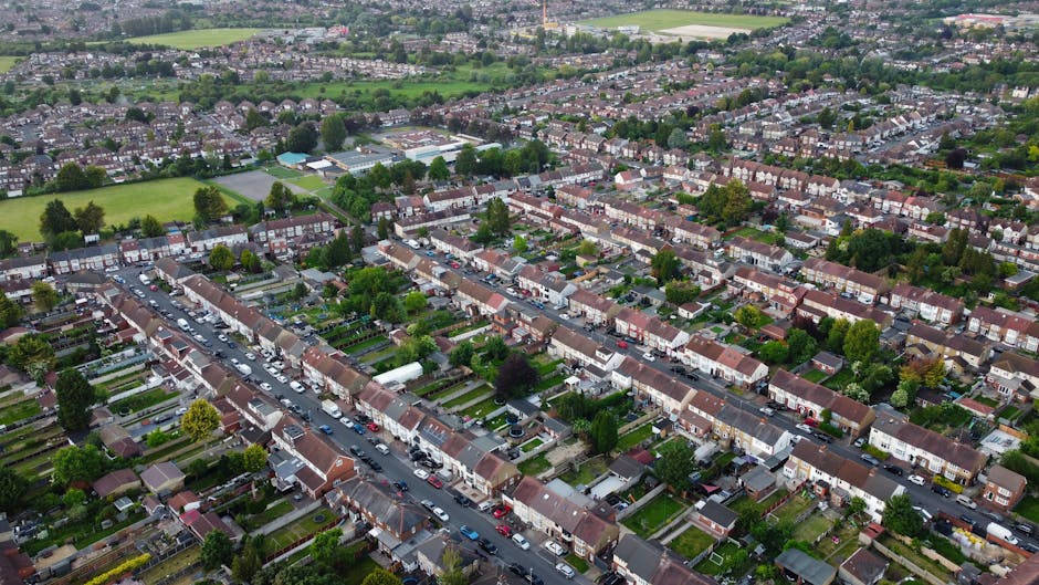 An aerial view of a suburb in South Tottenham showing a row of terraced residential houses with front gardens, parked cars along the street, and several trees lining the pavement. The image captures the quiet street scene during daylight, with the houses constructed from brick and featuring tiled roofs. Some houses have driveways or small yards, and there are visible footpaths and boundary fences. Beyond the row of houses, there are larger green fields and sports grounds, with additional residential areas extending into the distance. The overall scene reflects typical home relocation surroundings, with no moving activity visible but representing a neighbourhood where professional removals, packing, and furniture transport services by companies like Man with Van South Tottenham might operate.