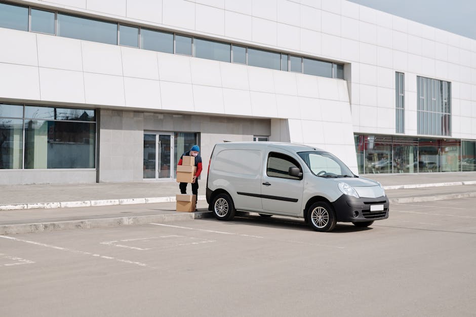 A man wearing a blue beanie, red jacket, and black trousers is standing outside a modern, white commercial building with large glass windows, loading several cardboard boxes into the back of a small grey van parked on a paved area. The boxes vary in size, and some are stacked next to him on the ground. The van is positioned close to the building's entrance, which features a glass door, and appears to be used for home relocation or furniture transport as part of a professional removals service. The background shows a clean, organized loading bay with marked parking spots and a clear, cloudy sky above. The scene reflects the process of packing and loading items efficiently in preparation for moving, with focus on the careful handling of boxes and use of the van for transportation. This visual is relevant for [PAGE_TITLE] on [DOMAIN], illustrating a typical loading process during house removals, managed by [COMPANY_NAME].