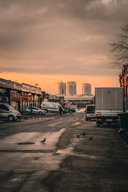 A wide view of a parking area in a commercial district during a sunset, with a cloudy sky casting an orange hue on the horizon. Several parked vehicles, including a small car, a large delivery van, and a cargo truck, are positioned along the pavement. The scene shows a person walking near the vehicles, possibly involved in loading or unloading items as part of a home relocation process. To the left, there are storefronts and shops with signage, indicating a retail area, while in the background, high-rise residential buildings are visible. The ground appears wet, reflecting some of the vehicle lights and the sky, with a few pigeons walking across the surface. This setting depicts a typical environment where professional removals services, such as those offered by Man with Van South Tottenham, coordinate the transport and packing of household furniture and boxes in preparation for moving home near Seven Sisters Road in N15, South Tottenham.