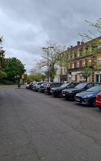 A row of parked cars, including sedans and hatchbacks in various colours, lines the side of a quiet residential street near Seven Sisters Road in N15, South Tottenham. Behind the vehicles, there is a multi-storey Victorian-style building with brickwork, large windows, and a small shopfront on the ground floor. A few trees with budding leaves are situated along the pavement, and the sky above is overcast with grey clouds, indicating typical weather conditions for an urban area. The scene suggests a location where a professional removals company, such as Man with Van South Tottenham, might facilitate home relocation or furniture transport, with the parked vehicles aligned along the curb ready for loading or unloading of boxes, furniture, and packing materials. The image captures the environment in which moving services would operate, with the street facilitating easy parking for transporting household items during a packing and moving process.
