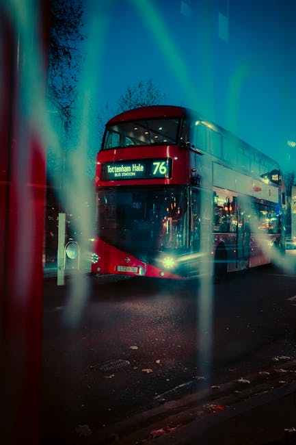 A double-decker bus displaying route number 76 and destination 'Tottenham Hale' parked on a street at night, illuminated by streetlights and the bus's headlights. The bus is red with a black front, and reflections of nearby buildings and lights are visible on its glazed upper deck. The image is taken through vertical, semi-transparent glass or plastic strips, creating a blurred, streaked foreground effect, with the bus positioned centrally within the scene. The dark sky and illuminated surroundings highlight the urban environment typical of a home relocation or moving process in South Tottenham, supported by local transport infrastructure. The scene captures the typical setting where furniture transport and moving logistics are coordinated, with the bus ready for passenger journeys or through traffic, as part of the area’s daily movement flow.
