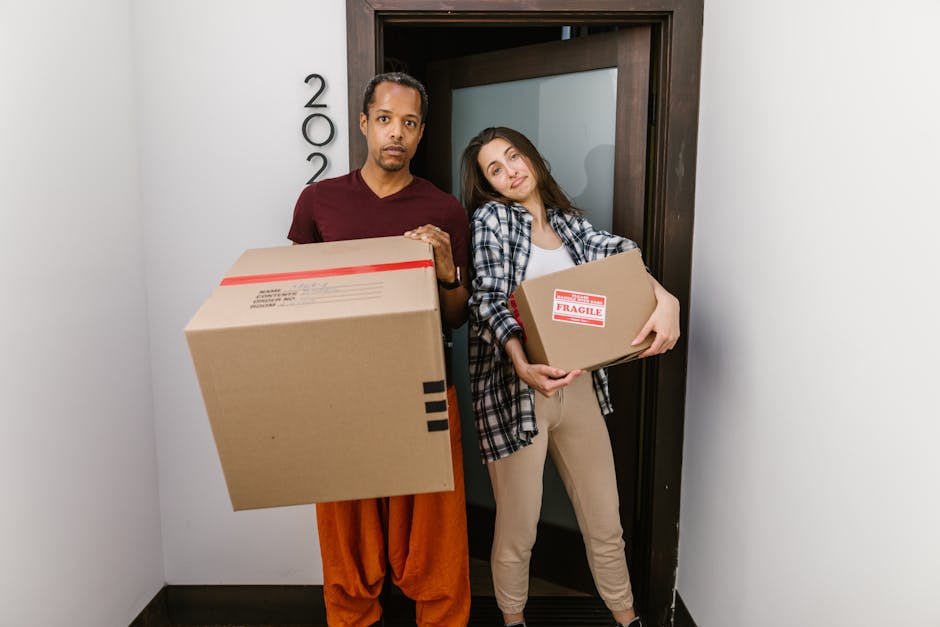 A man and a woman stand inside a residential property near a doorway, holding cardboard boxes prepared for a home relocation. The man is wearing an orange pair of trousers and a maroon shirt, carrying a large box with black markings. The woman, dressed in beige trousers and a plaid shirt, is holding a smaller box with a red 'Fragile' label. Behind them, a dark wooden door with a frosted glass panel is partially open, revealing a hallway or another room. The scene takes place indoors, with neutral-colored walls and a dark floor, and is set in a professional moving context, where [COMPANY_NAME], Man with Van South Tottenham, is assisting with packing and furniture transport as part of the removal and moving services aimed at ensuring safe and efficient house moving logistics.