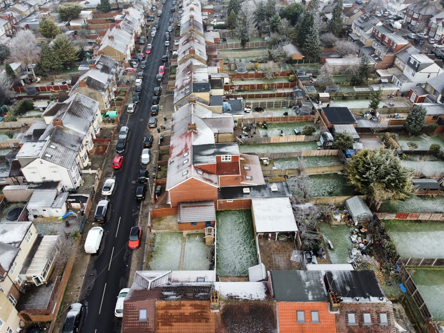 An aerial view of a residential street in South Tottenham during winter, showing houses with snow on rooftops and gardens. The street is lined with parked cars on both sides, with some cars partially covered in snow. The houses vary in style, with brick facades, pitched roofs, and some flat-roofed structures. The backyards are enclosed with wooden fences, with some gardens featuring trees, sheds, and outdoor furniture. The road appears to be a narrow, one-way street with minimal traffic. The image depicts a calm neighborhood scene with natural daylight and overcast sky. This setting illustrates the typical environment where house removals and furniture transport services might be carried out, reflecting the residential layout and logistics involved in home relocation. Occasionally, Man with Van South Tottenham provides professional removals in similar neighborhoods, supporting efficient packing and moving processes across these homes.