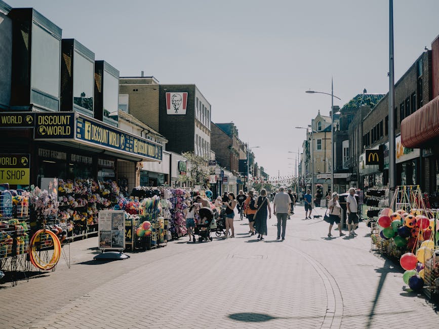 A busy retail street scene in South Tottenham featuring various small shops and vendors selling flowers, balloons, and other merchandise on both sides of the pavement. The shops include a discount store, a fast-food outlet with a prominent McDonald's sign, and other storefronts displaying colorful displays and signage. Pedestrians, some pushing strollers, are walking along the wide, paved pedestrian zone under bright, clear weather. The street is lined with tall buildings, with a few street lamps and signage visible, and the scene suggests a lively commercial area typically involved in home relocation logistics, such as loading and unloading goods during moving or packing activities. This setting aligns with local moving services provided by Man with Van South Tottenham, who may facilitate furniture transport or packing and moving in this area.